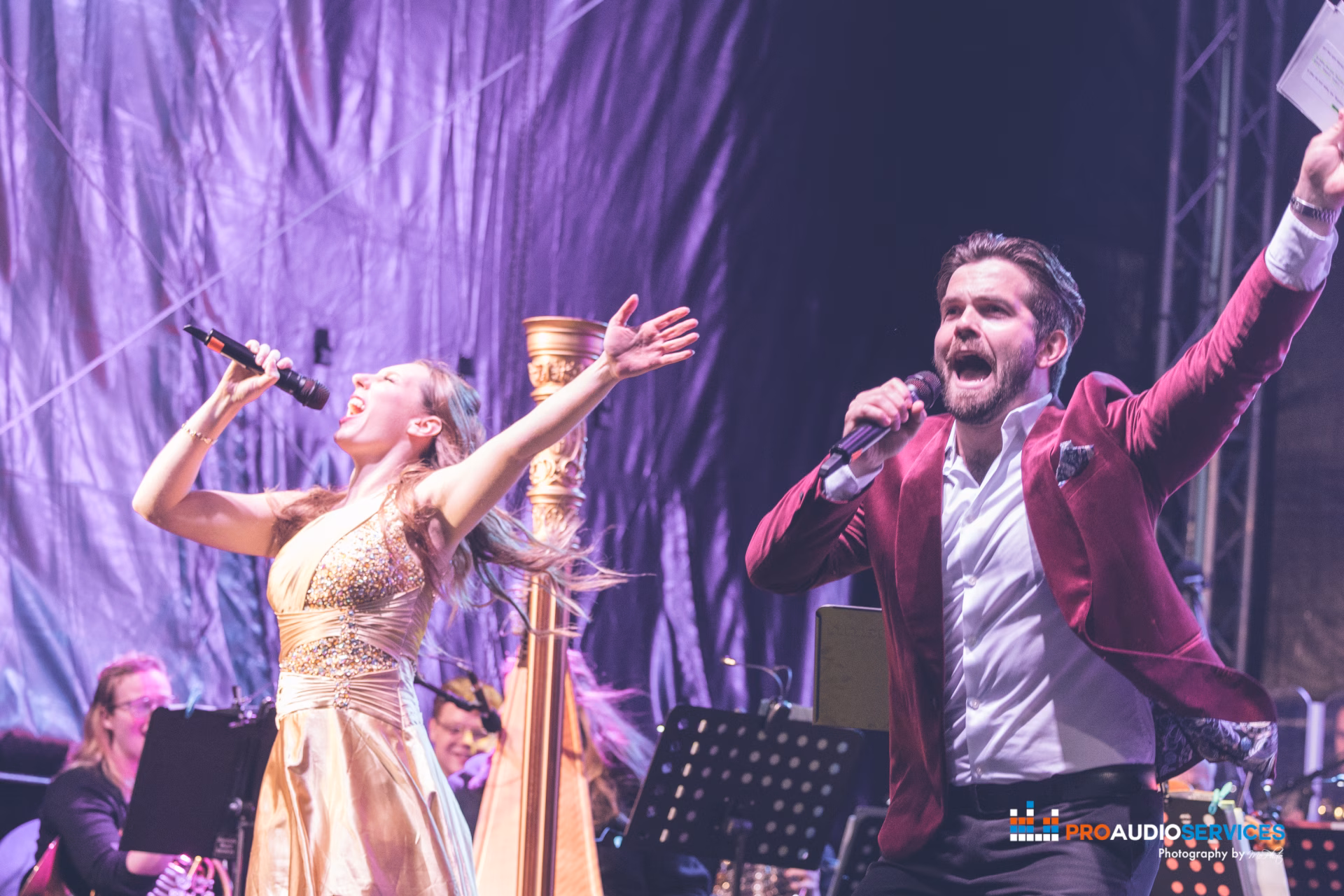 Two vocalists performing on stage with a harpist behind, under purple drapes