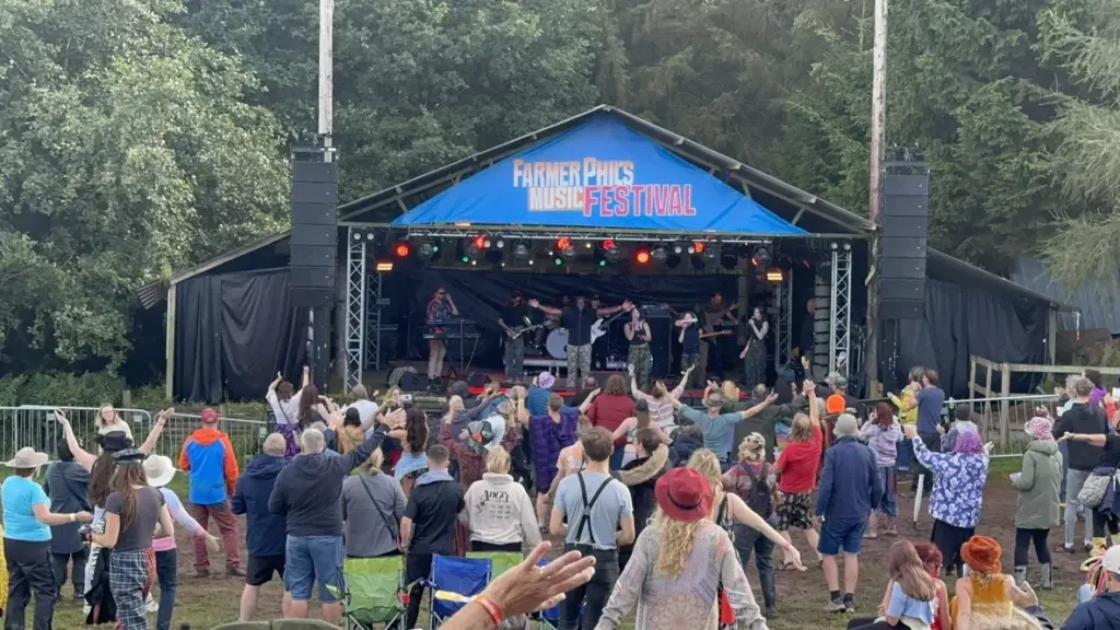 Festival crowd in front of a blue-roofed stage among trees