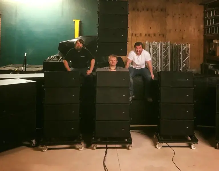 Three people sitting on top of stacked line array speaker cabinets in a warehouse