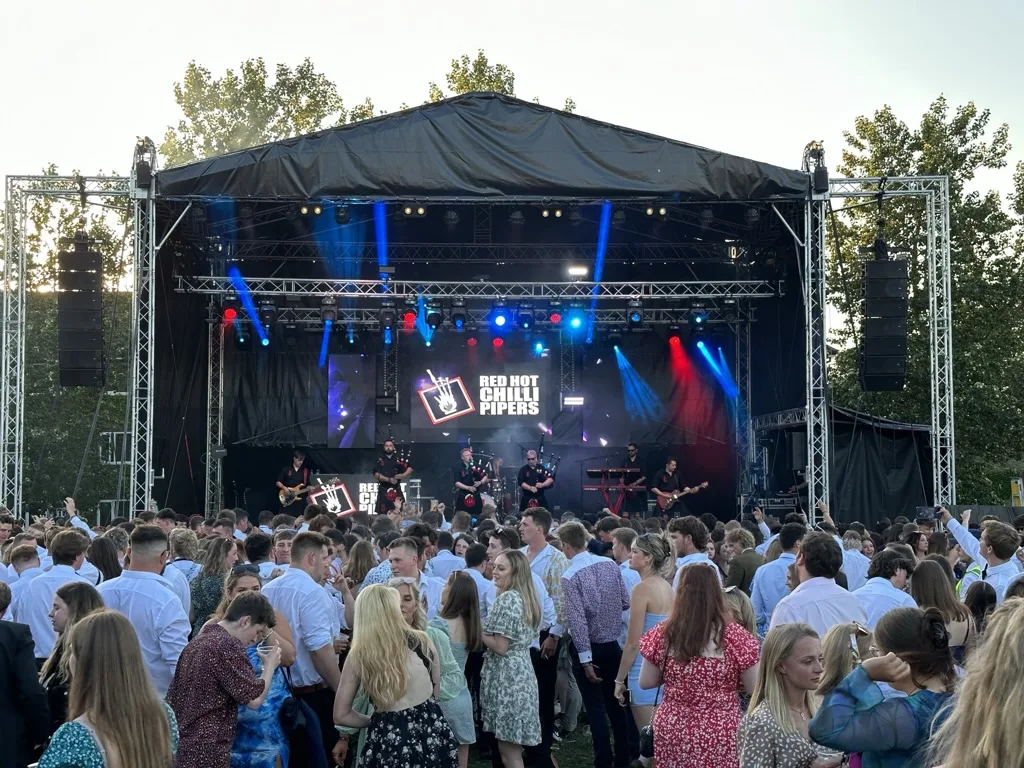 Large crowd in front of a covered festival stage with lighting rigs, filmed in evening light