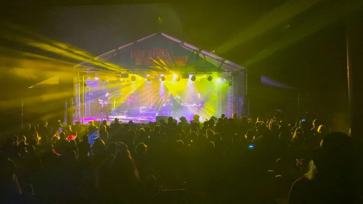 An audience watching a band play on a stage at night with yellow and pink lights coming out from behind the bnad