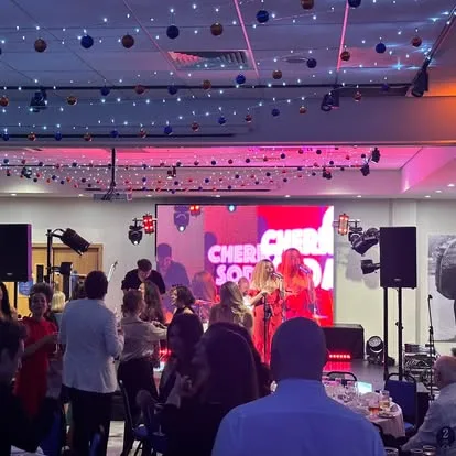 People dancing in a room infront of a platform. On the platform is a lady singing. There is a video wall behind her with her showing on the screen. There are lights accross the ceiling.