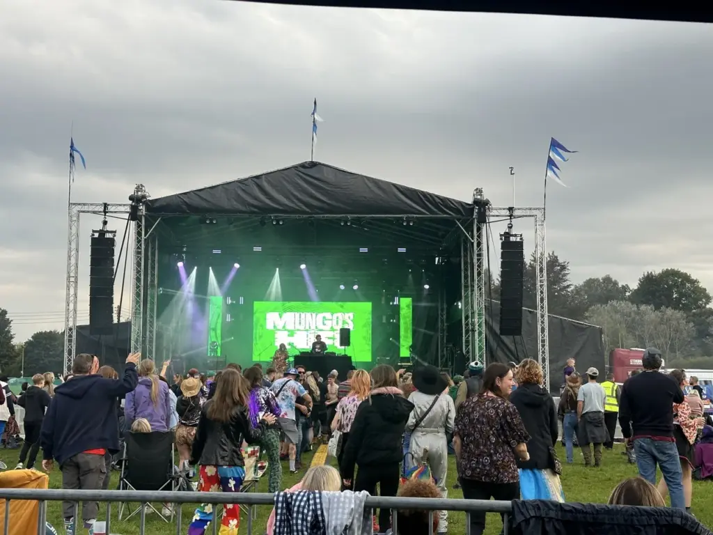 Green-lit stage in a field with a crowd in front under a cloudy sky at Heal Festival