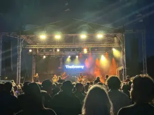Large crowd watching band (The Enemy) on dtage. There is a vidoe wall nehind the band displaying the band's name and yellow and orange lights shine onto the band and out into the audience