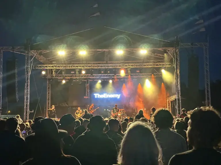 Large crowd watching band (The Enemy) on dtage. There is a vidoe wall nehind the band displaying the band's name and yellow and orange lights shine onto the band and out into the audience
