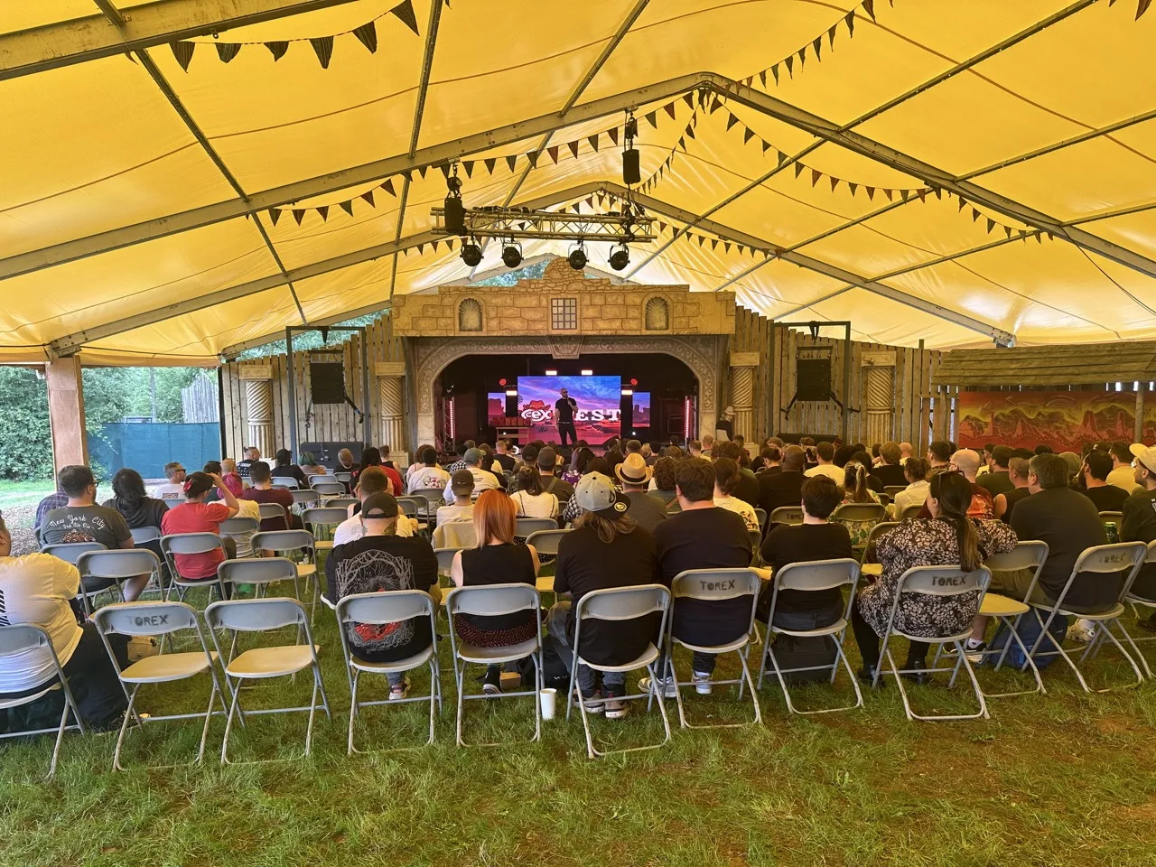 An audience sat on chairs in a marquee listening to a man talk on a stage with the CeX logo showing on a video wall behind him