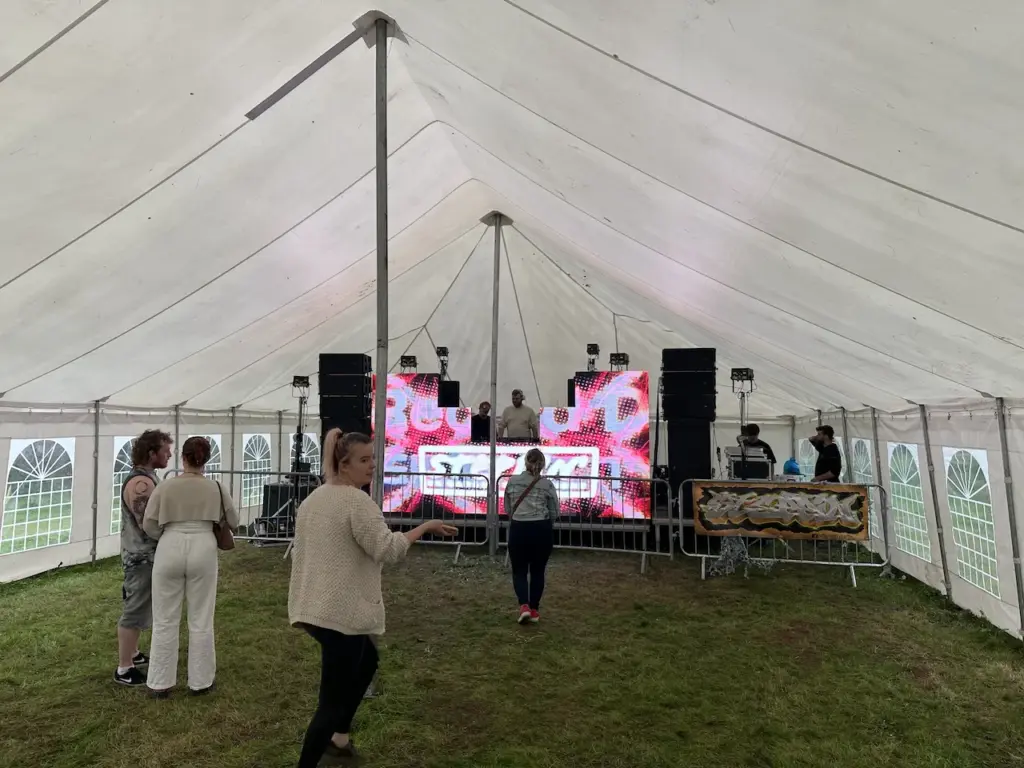 A couple of people dancing in a marquee infront of a DJ playing dance music with a video wall around him showing some colorful patterns