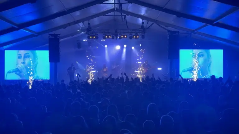 Large crowd dancing to a band on stage. There is a woman singing at the front of the stage and her face is shown on 2 video walls, one each side of the stage.