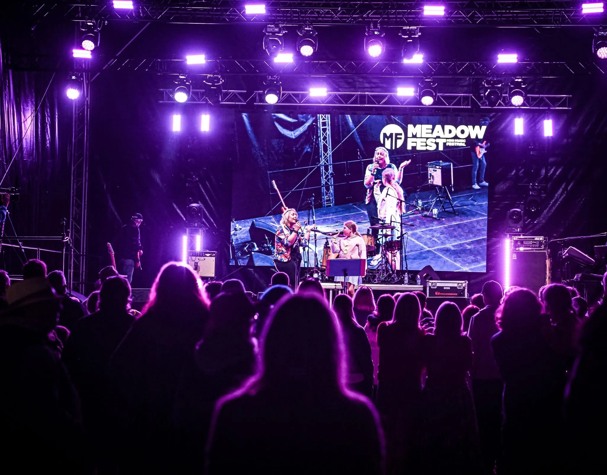 Crowd silhouetted by purple light shinning out from satge. A lady sings with her daughter on a stage and the image is showing on the video wall behind them.