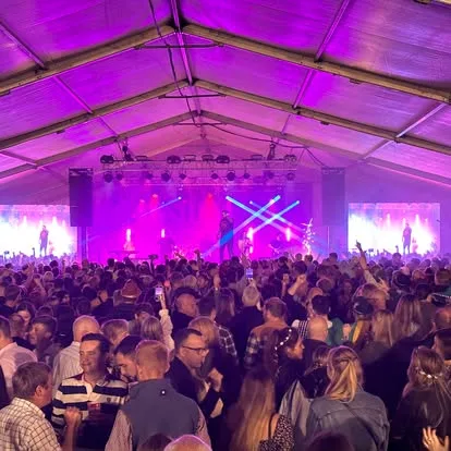 A large crowd of people standing in a marquee watching a band play on a stage. There are speakers and a video wall on each sude of the stage and there are pink and blue lights shinning both on the band and the audience.