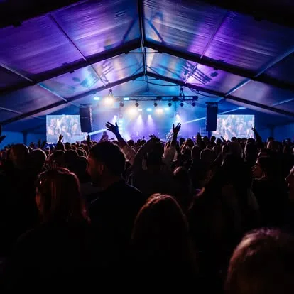 Stage inside a marquee with blue and magenta lighting, LED screens either side and the crowd at the front