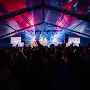 Large audience dancing in front of a stage where a band is playing. There is a video wall each side of the stage and there are red and blue lights shinning out into the audience.