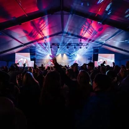 Large audience dancing in front of a stage where a band is playing. There is a video wall each side of the stage and there are red and blue lights shinning out into the audience.