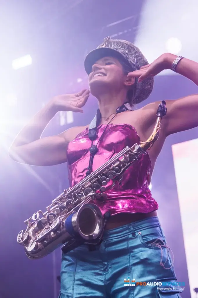 Saxophonist in a sequinned silver top and cap mid-performance, lit in pink and purple