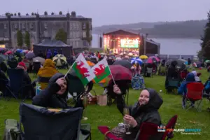 Large statley home with a river behind and stage next to it. People with umbrellas watching an orchestra play on the stage waving the Welsh flag.