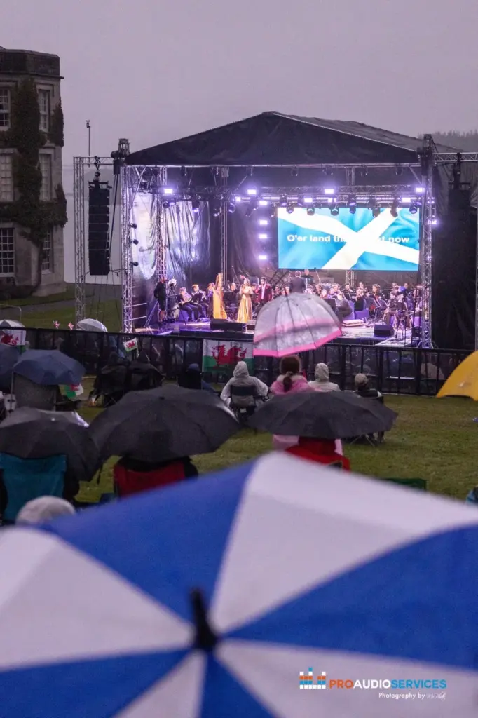 Outdoor stage with a flag on its LED screen, seen over audience umbrellas in the rain