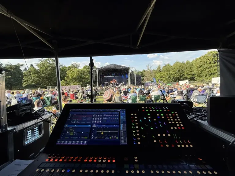 A sound desk at the front of the front of the picture with a field of people watching an orchestra play on a stage. The sun is shinning and there are trees around the field.