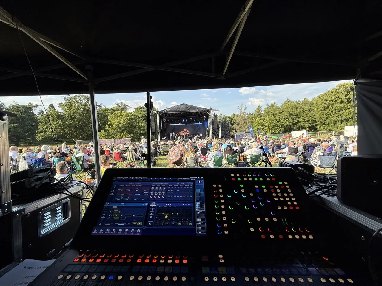 A sound desk at the front of the front of the picture with a field of people watching an orchestra play on a stage. The sun is shinning and there are trees around the field.