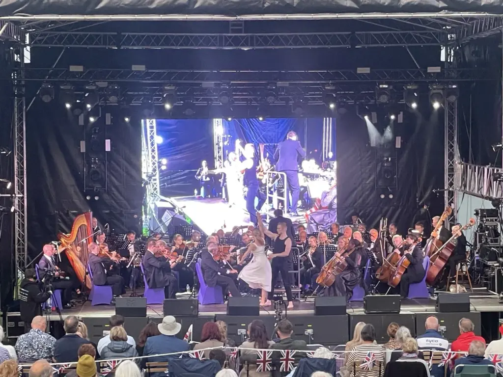 Wide shot of the orchestra on stage with an LED screen and a dancer, audience seated in the foreground