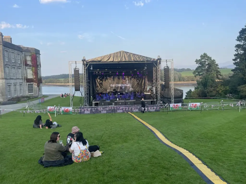Audience on the grass in front of a distant stage at Proms in the Park, Plas Newydd, with the water beyond