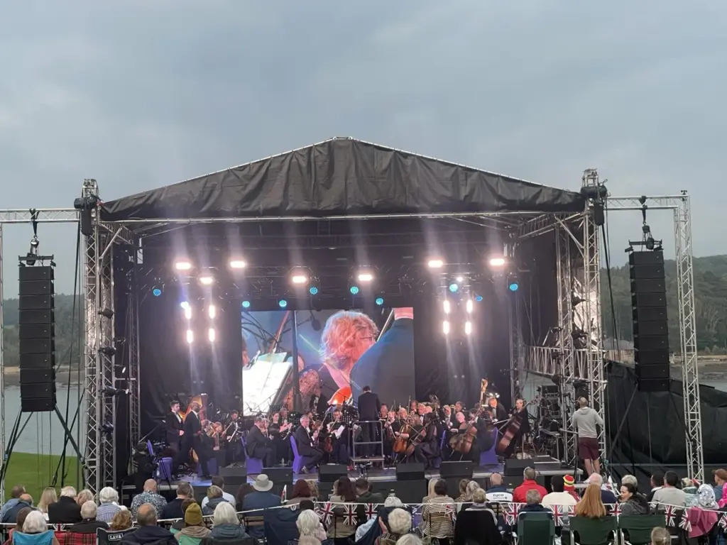 Orchestra on an outdoor stage with a large LED screen and speaker stacks either side, audience seated in front