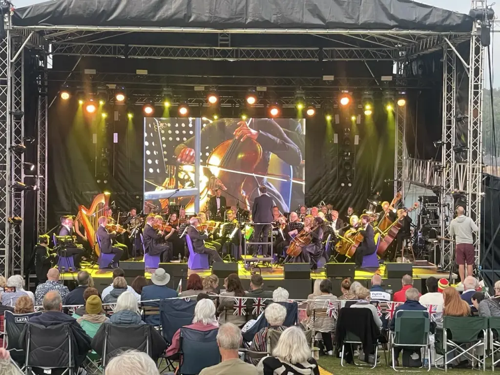 Orchestra performing on a roofed outdoor stage with an LED screen and warm stage lighting