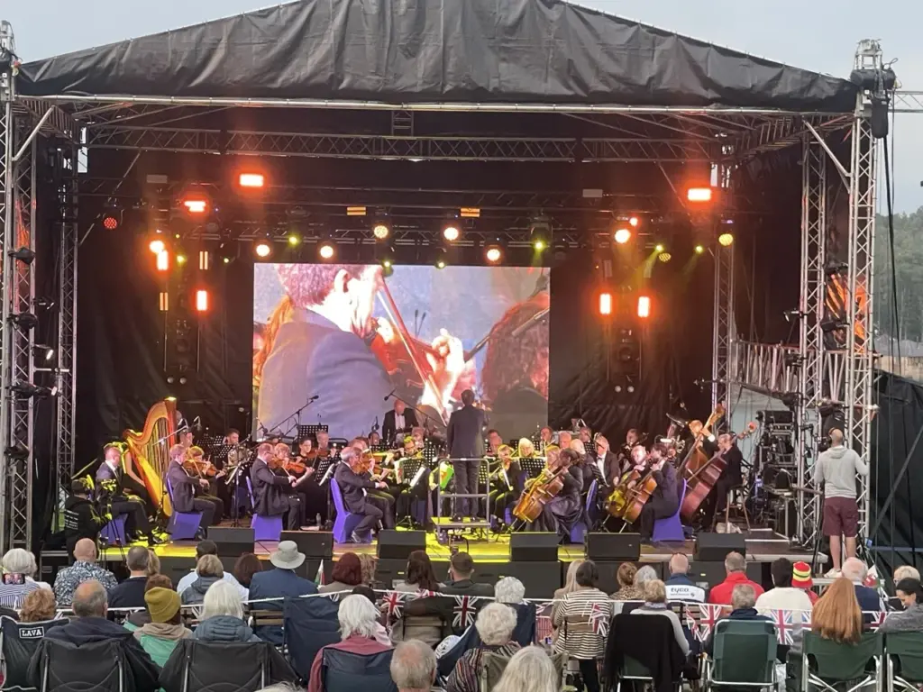 Orchestra and conductor on stage under a black canopy with an LED screen and amber lighting