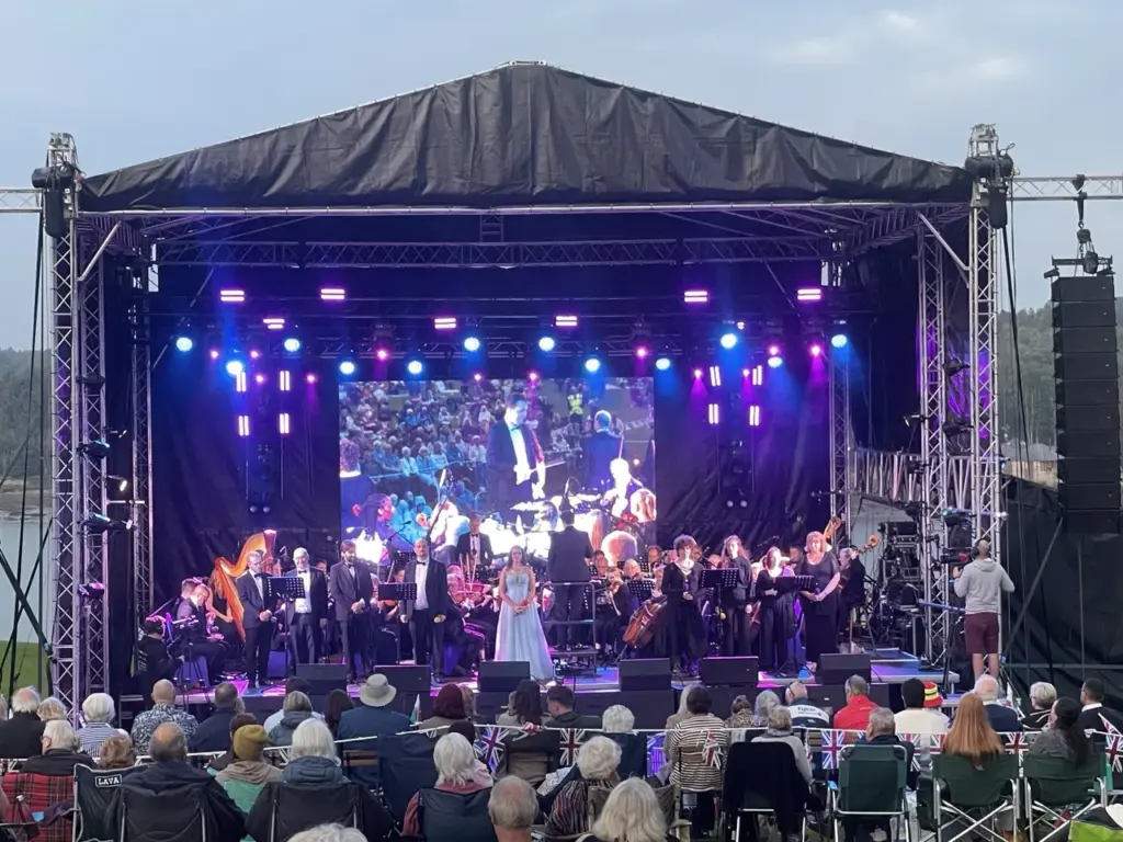 Orchestra playing on a covered outdoor stage with LED screens and stage lighting at Proms in the Park, Plas Newydd