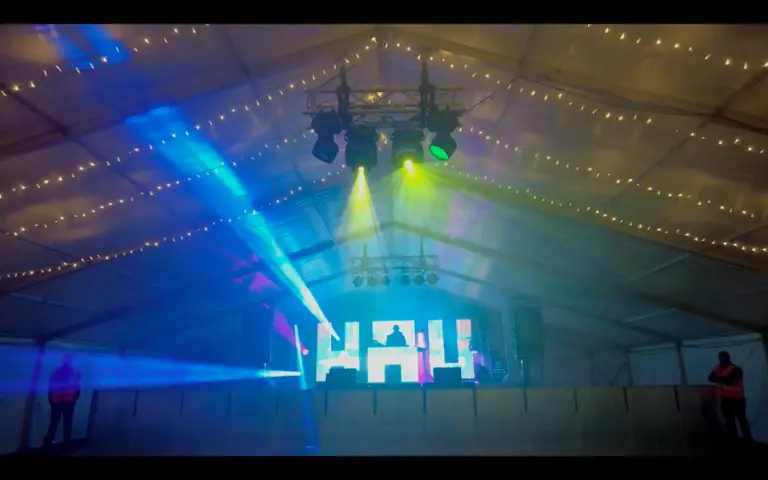 A stage in a marquee with a video wall in the shape of HAU. There is a DJ playing on the stage and some lights shine out around him.