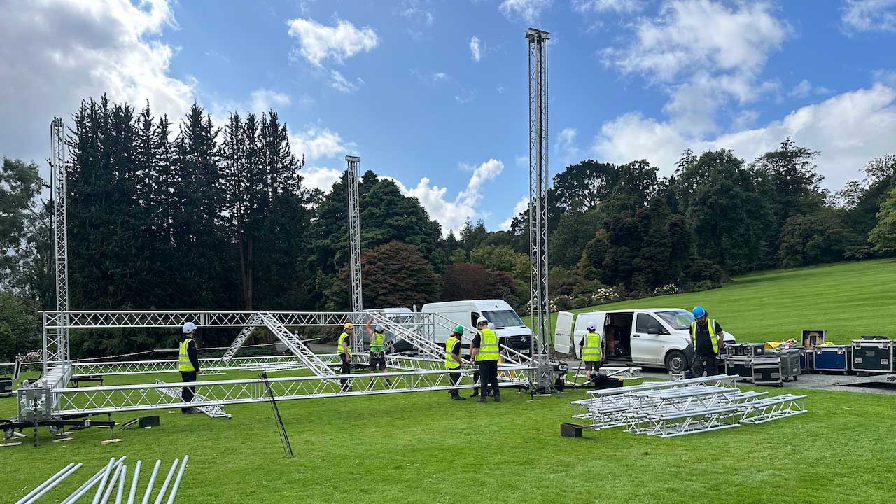 Stage build team working with truss framework laid out on grass. Riggers wearing hard hats, high-visibility vests and climbing equipment.