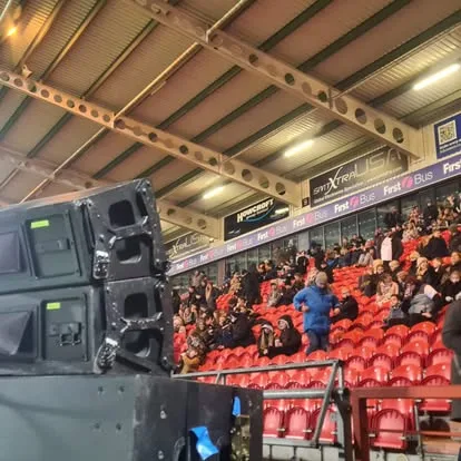 Stacked line array speakers pointing at the stand, with spectators in red seats beyond