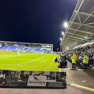 The edge of a football pitch. An audience sitting and looking towards the pitch with some speakers directed towards the audience.