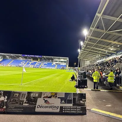 The edge of a football pitch. An audience sitting and looking towards the pitch with some speakers directed towards the audience.