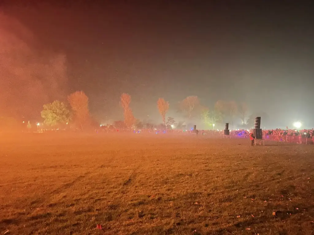 Orange-lit smoky field at dusk with trees on the horizon during a firework show