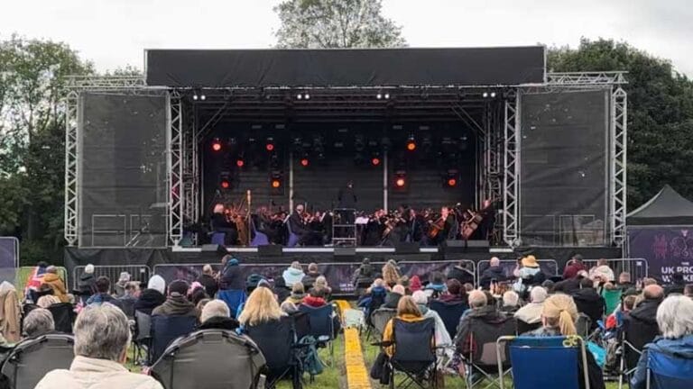 People sat on chairs outside watching a live orchestra play on a stage with orange lighting behind.