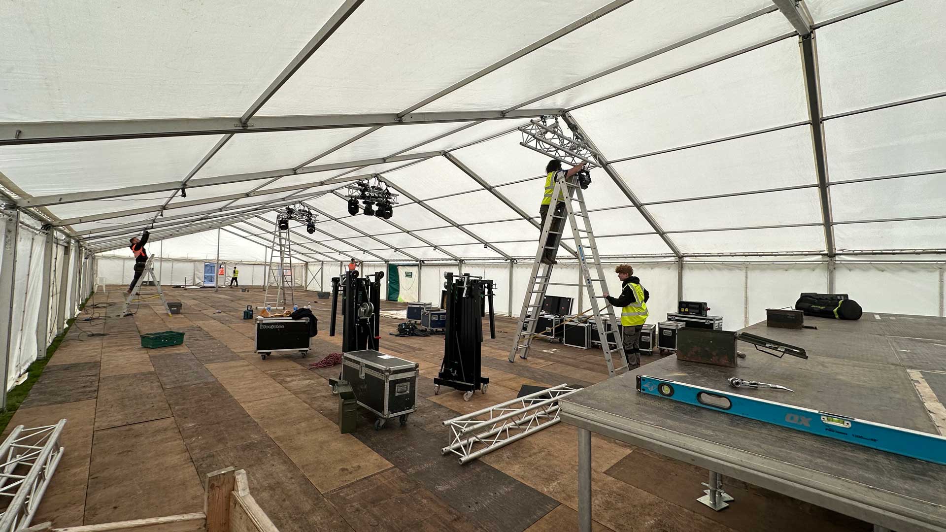 Crew in hi-vis vests rigging lights from ladders inside a large white marquee, with flight cases on a raised wooden stage deck below