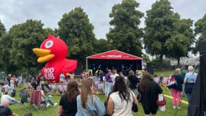 People outside watching a band perform on a stage with a huge inflatable duck beside it and some trees behind it.
