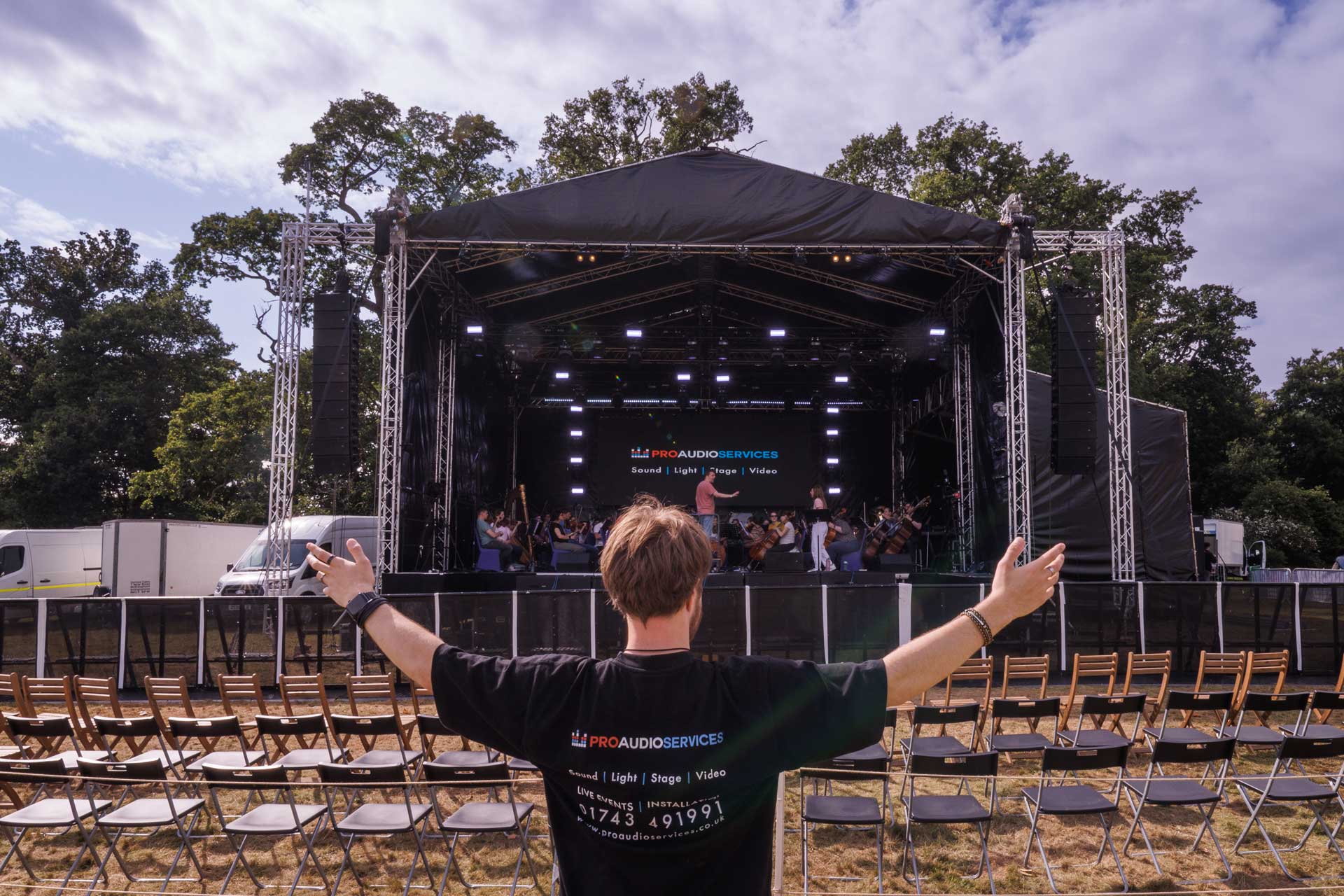 Person stood in front of larger concert sound system and stage with orchestra rehearsing