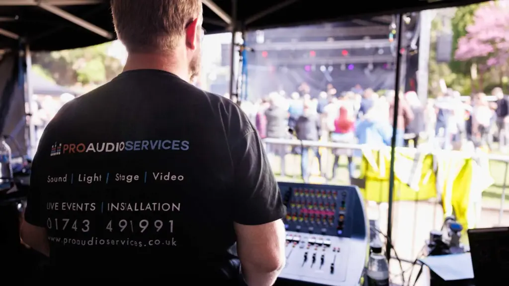 Looking over the shoulder of a sound engineer in front of mixing console looking towards a stage with audience watching.
