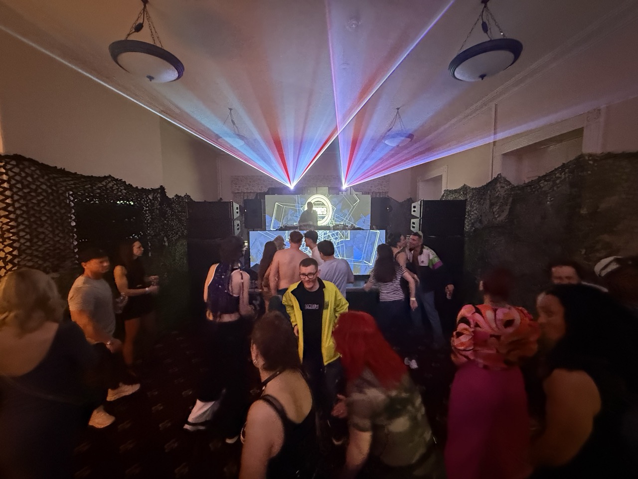 People dancing in a room with a DJ playing. There is a video wall in front and behind the DJ and lasers firing over his head.