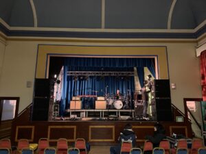 Astage in a hall set-up with speakers each side, drums on a drum riser in the middle.