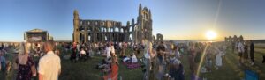 People sat and walking on the grass field in front of Whotby Abbey on a sunny evening.