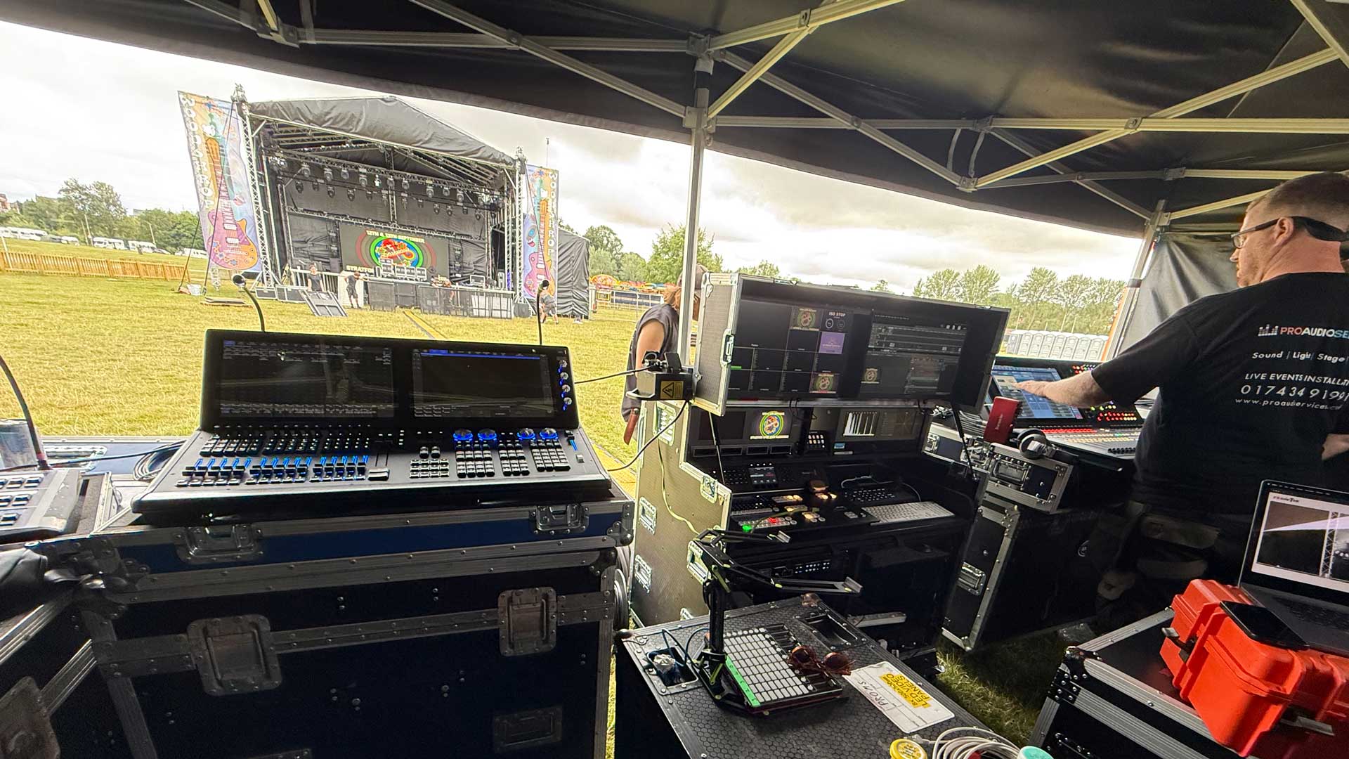 Front-of-house mixing desk and flight cases under a canopy, with a festival stage in the distance