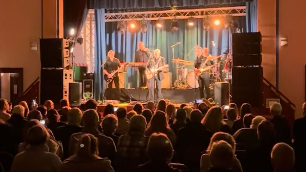 Band on stage in a venue under warm lighting with the audience in the foreground