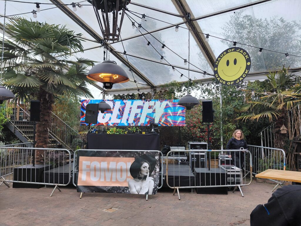 DJ booth with a branded front and crowd barriers inside a glass-roofed marquee