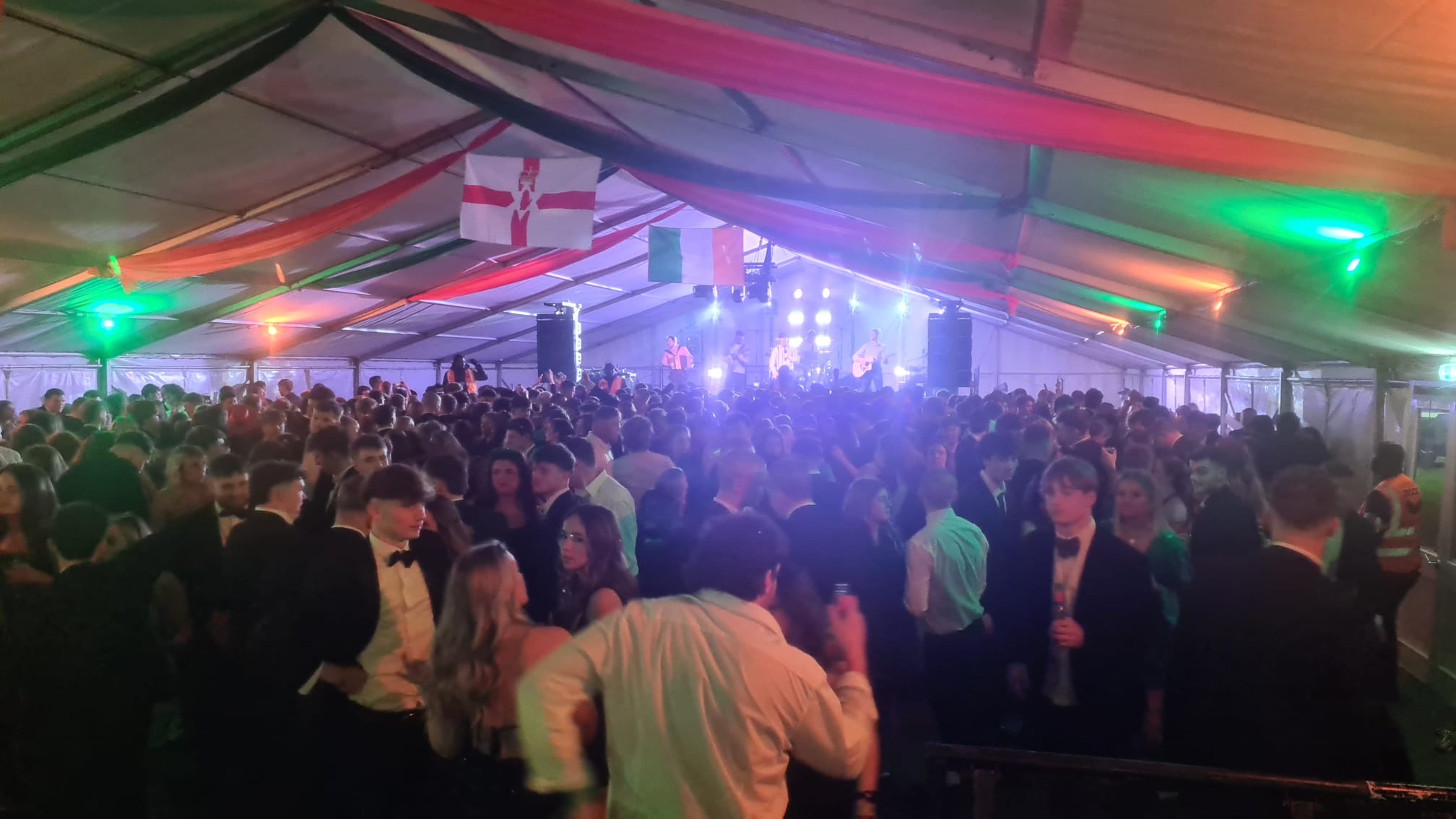 Amarquee full of young people dancing infront of a stage woth a band playing it and lighting shinning pout from behind them.