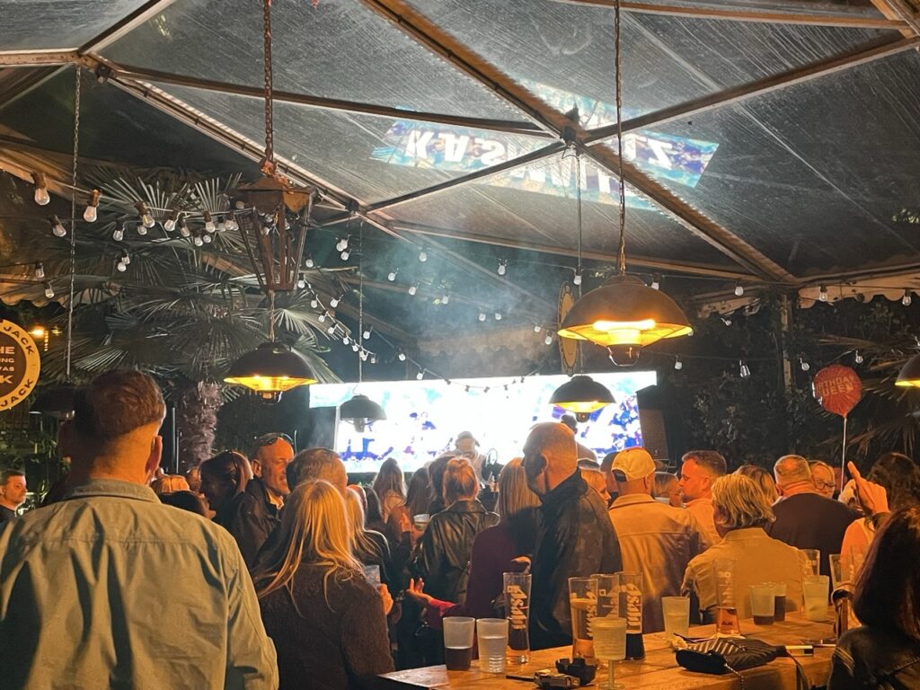 People gathered in a glass-roofed marquee with a video screen and hanging lights
