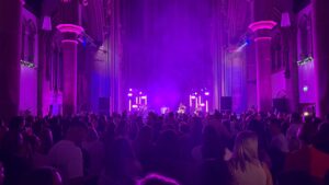 Monastery church space with band playing at a stage at the end of a church in front of a packed full audience. Pink and purple hue lighting creating atmostphere.