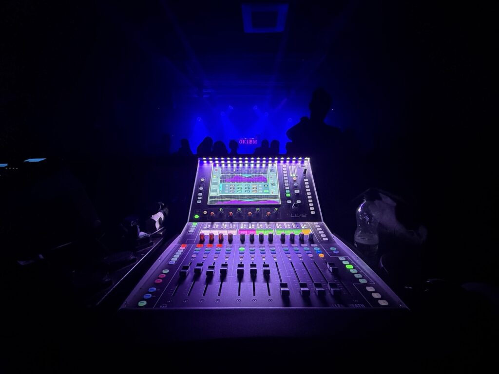 Digital mixing console lit in purple in a dark room, seen from behind
