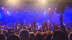 A band playing on a stage with an audience standing in front with their arms held up in the air.